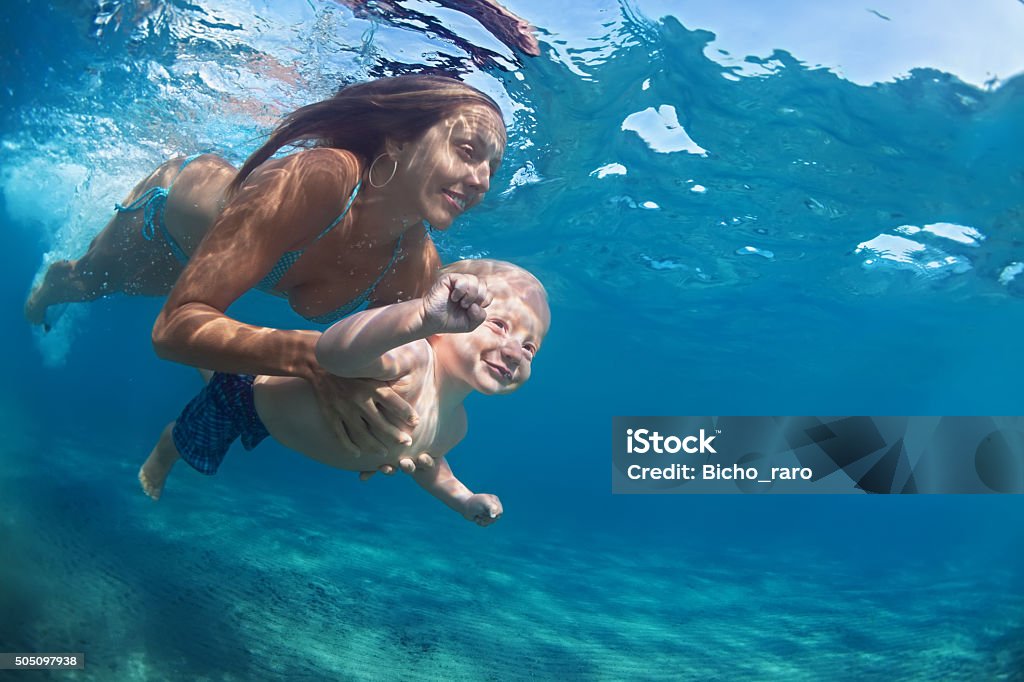 Mother with child swim underwater in blue beach pool Happy family - mother hold in hands baby girl and dive down with fun in tropical beach sea pool. Active parent lifestyle, people water sport activity, underwater swimming lesson on vacation with child Child Stock Photo