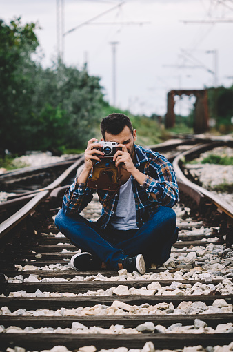 Young hipster guy tourist with camera and bag photographing on the railroad. Vintage Instagram style effect, soft and selective focus, grain texture visible on maximum size