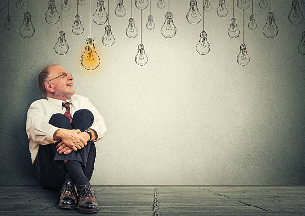 Man sitting on floor looking up at light idea bulb Portrait thinking elderly man in glasses sitting on a floor looking up with light idea bulb above head isolated on gray wall background. man with a light bulb over his head stock pictures, royalty-free photos & images