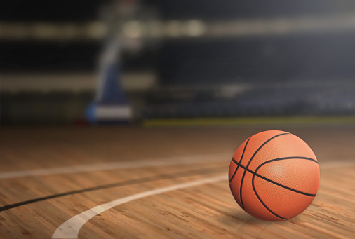 A shot of a female teenage athlete sitting on the floor during half time of a basketball game. She is smiling and looking into the camera. She is socialising with her team mates with a basketball by her side.