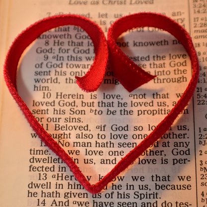 Bible page of the First Epistle of John 4:10, with red heart layed over the text. Christian Valentine's Day background. Red ribbon in shape of heart.