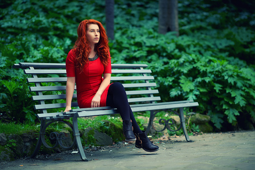 young red hair woman in red dress sitting in park on a bench and relaxing,spring season.green trees around her.