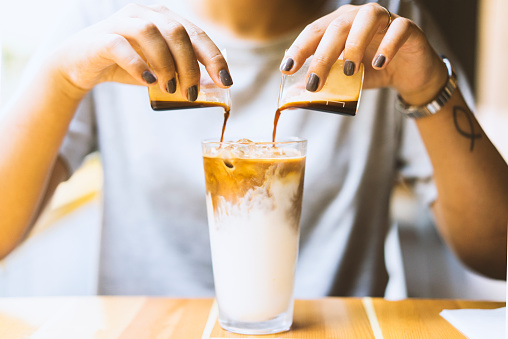 Woman pouring espresso shots into a glass of milk with two hands, preparing latte.