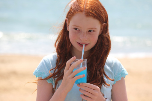 Photo showing a girl with long red hair and a summer T-shirt, drinking a slushie ice drink with a straw.  The girl is pictured in the full sunshine, standing on a beach while she drinks her blue, raspberry flavoured ice / frozen drink, with the lapping waves of the sea sparkling in the background.