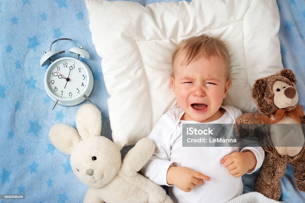 One year old baby with alarm clock One year old baby lying in bed with alarm clock and crying Baby - Human Age Stock Photo