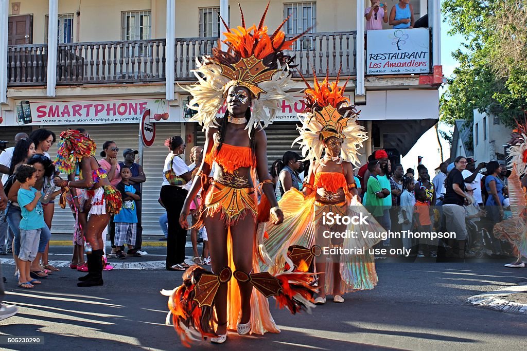 Marigot Carnival 2015 - 2 Marigot, Saint Martin (French part) - February 15th, 2015: Two participants on parade in Rue de la Liberté, Marigot during the Marigot Carnival or "Carnaval de Marigot", an annual event / celebration in Saint-Martin. They wear bright orange colored costumes decorated with flamboyant feathers on the headdress. People watch the parade from the sidewalk. Another participant is interacting with the crowd of spectators. Antilles Stock Photo