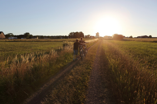 Mother and son walking with bicycles on a country road surrounded by barley fields at sunset.