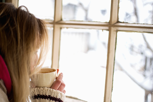 Closeup of woman having a cup of tea or coffee on snowy day. She's sitting in a cafe and looking through window. Wearing knitted cardigan and scarf and enjoying winter in warm and cosy place. Low angle shot.