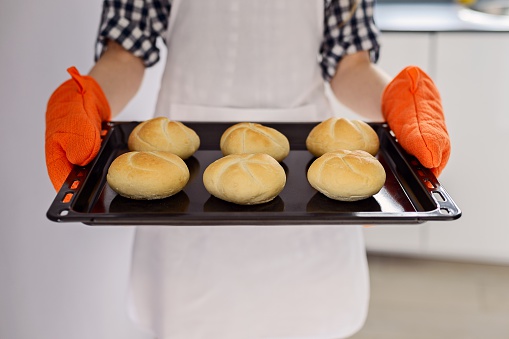 woman holding a baking tray with freshly baked bread rolls. Orange kitchen gloves on hands