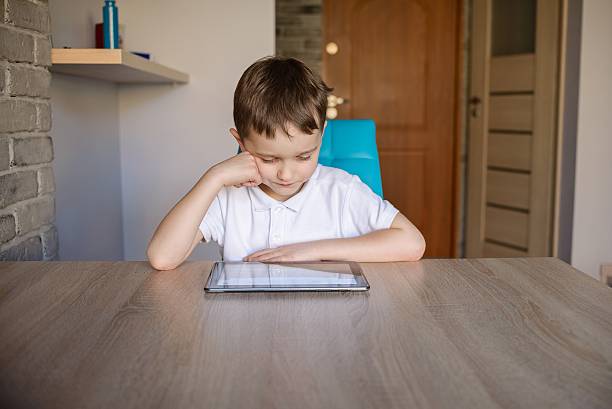 chico usando tableta mientras se distiende en la mesa de comedor. - video game child handheld video game little boys fotografías e imágenes de stock