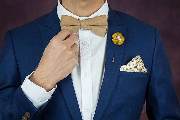 Man in blue suit with coffee cream bowtie color, flower brooch, and dot pattern handkerchief, close up, adjusting bowtie