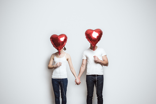 A young couple wearing white shirts and blue jeans hold red heart shaped balloons to celebrate the Valentine holiday.  They hold hands, their faces obscured by the balloons.  Horizontal image white background and copy space.