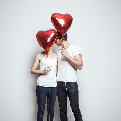 A young couple wearing white shirts and blue jeans kiss, their faces partially obscured by red heart shaped balloons to celebrate the Valentine holiday.  Square crop with white background and copy space.