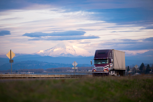 Dark professional semi truck with trailer transporting commercial goods on the scenic road with a metal security fence on the background of snow-capped Mount Hood and a dark cloudy sky.