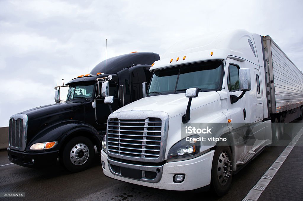 Two contrast modern semi truck different models black and white Two contrasting shiny modern black and white big rigs semi trucks with a trailers and a high sleeper cab for truckers relaxing on truck stop move side by side along the interstate highway carrying commercial goods. Two Objects Stock Photo