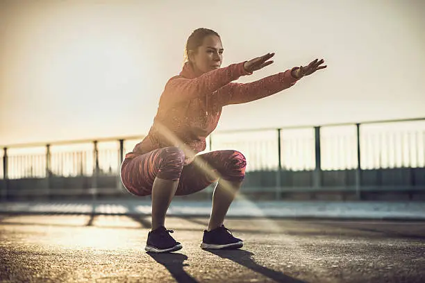 Athletic woman exercising outdoors at sunset and doing squats.