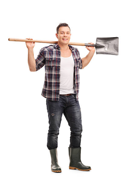 A happy male farmer wearing a straw hat and standing in a well-maintained garden holding a wooden tool. The lush greenery and clear sky create a serene and productive ambiance.