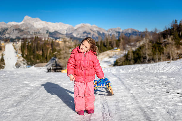 Girl is pilling snow sledge in ski resort  wearing winter clothes. High mountain ski resort, vacation, holiday and sunny day mountain peaks.