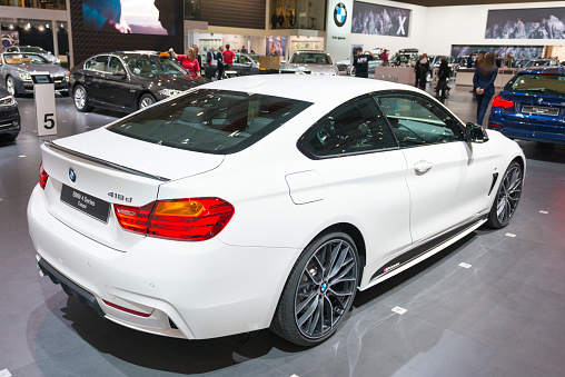 Brussels, Belgium - Januari 12, 2016: White BMW 4 series coupe (model F32) rear view. This white version on display is the 418d, fitted with a diesel engine. The car is on display during the 2016 Brussels Motor Show. The car is displayed on a motor show stand, with lights reflecting off of the body. There are people looking around and other cars on display in the background.