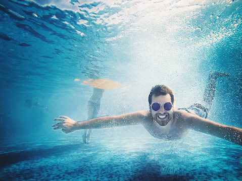 Closeup of smiling man diving in a swimming pool. He's moving towards camera very close to the bottom. Wearing sunglasses.