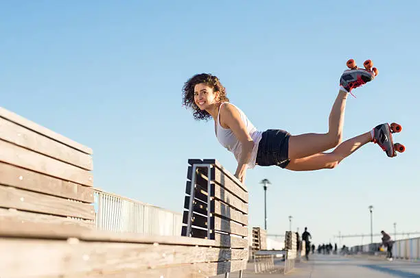 Happy young joyful woman doing a balancing act wearing rollerskates. Young woman in skates jumping with the support of a bench. Beautiful young happy woman wearing roller blades doing a stunt.