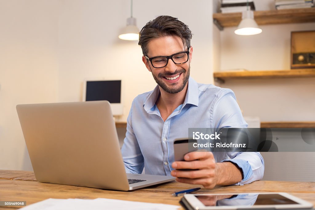 Happy man using smartphone Young happy businessman smiling while reading his smartphone. Portrait of smiling business man reading message with smartphone in office. Man working at his desk at office. Men Stock Photo