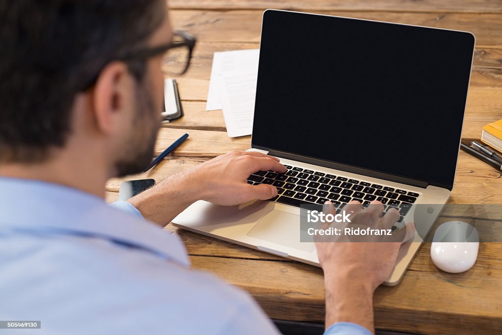 Man working on laptop Back view of businessman sitting in front of laptop screen. Man typing on a modern laptop in an office. Young student typing on computer sitting at wooden table. Laptop Stock Photo