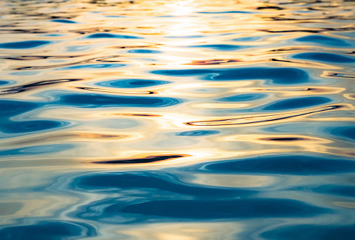 A colourful background texture of soft, calm waves on the surface of a body of tranquil water, photographed outdoors at sunset.