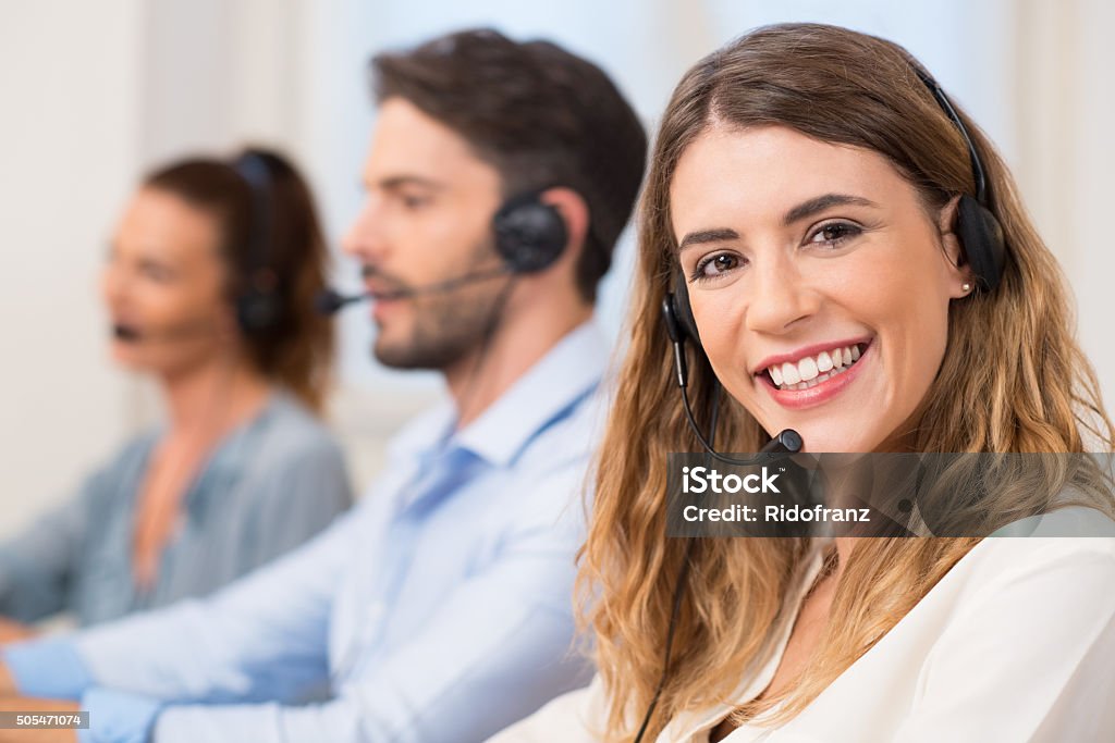 Woman at call centre Smiling female call centre operator doing her job with a headset while looking at the camera. Portrait of happy woman in a call center smiling and working. Portrait of happy smiling female customer support phone operator at workplace. Call Center Stock Photo