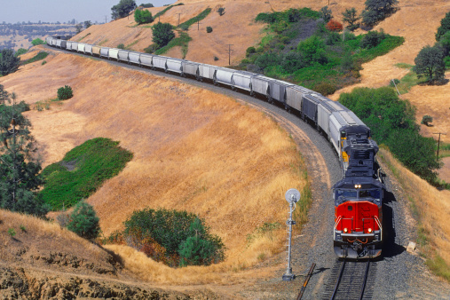 A heavily loaded grain train hauled by two diesel-electric locomotives winds its way round a curving hillside embankment on the way from farm to port.  Horizontal, copy space, numbers and logos removed.