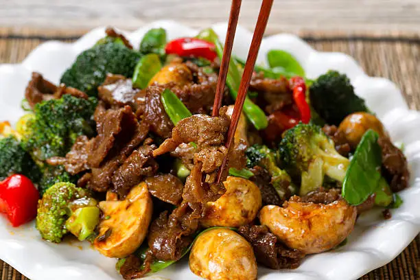 Close up view of tender beef slices, mushrooms, broccoli, peppers and peas in white plate. Selective focus on single piece held by chopsticks.