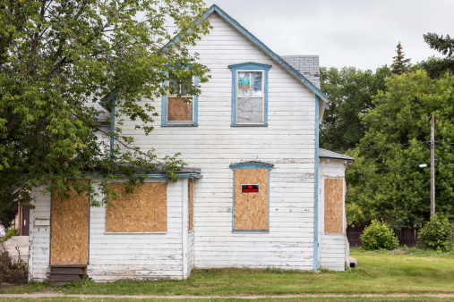 an old white condemned two-story house with windows boarded up and a sign saying 