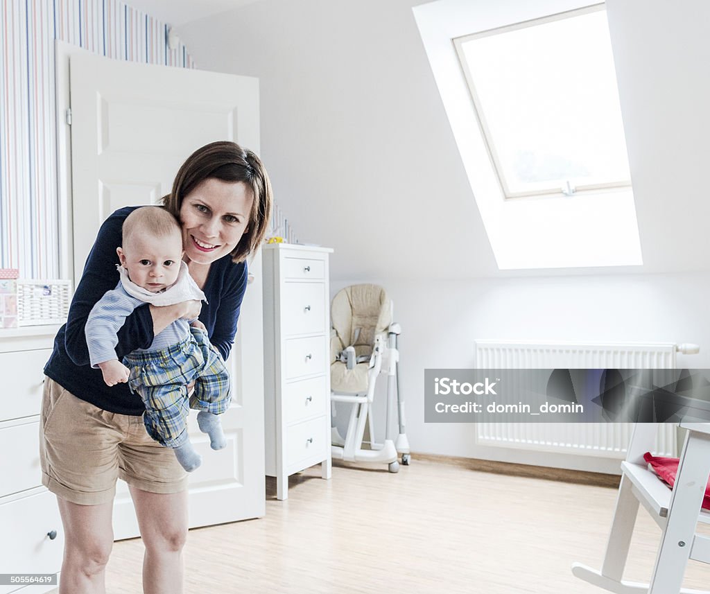 Smiling mother holding little baby boy on hands, baby's room Say hello to your Dad! Mother with her little son on hands smiling. Little baby's room, home interior with white furnitures, red and blue striped wallpaper. 0-11 Months Stock Photo