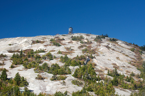 Close view of sloping, granite bedrock on the summit of Mt. Cardigan, with firetower, pine trees, and blue sky, summertime near Grafton in northern New Hampshire.