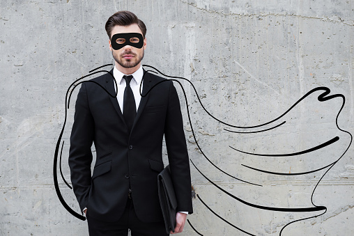 Confident young businessman wearing a drawn cape and mask while standing against concrete wall