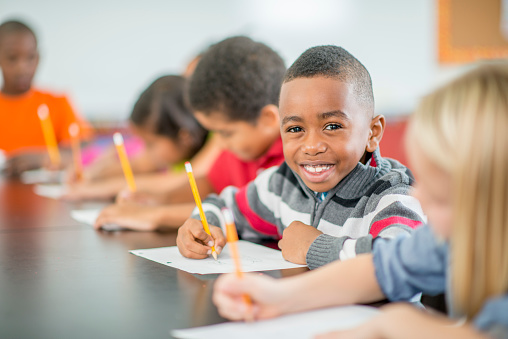 A multi-ethnic group of elementary age children are sitting at their desk and are taking a test in class. One boy is smiling and looking at the camera.