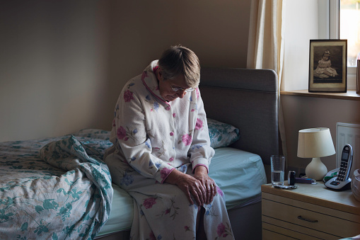 A shot of a senior woman sitting on the side of her bed with her head down looking sad. She looks like she has just woken up as is dressed in nightwear.