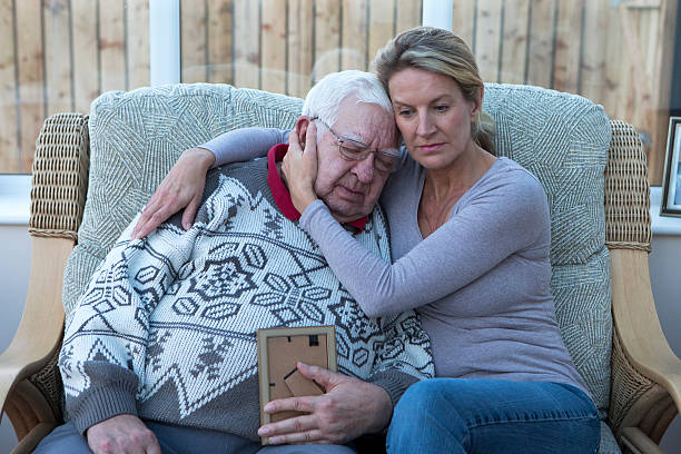 Losing a Loved One A shot of a senior man sitting on the sofa with his daughter, looking at a photograph. He is cuddling his daughter, looking sad and holding the photo. father daughter 80s stock pictures, royalty-free photos & images