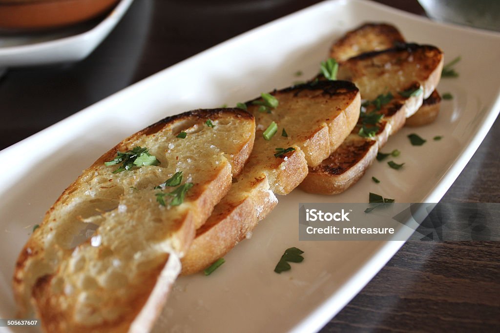 Image of garlic bread slices, topped with herbs and toasted Photo showing slices of grilled garlic bread, toasted with fresh herbs, including parsley.  The garlic bread was served on a long white plate in a Spanish restaurant, as part of a 'tapas' meal. Appetizer Stock Photo