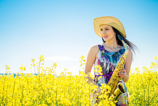 Young beautiful woman with saxophone in yellow rapeseed field and blue sky
