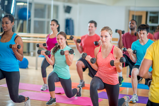 A multi-ethnic group of young adults are working out together in a aerobic fitness class at the gym. They are doing forward lunges with weights on their fitness mats.