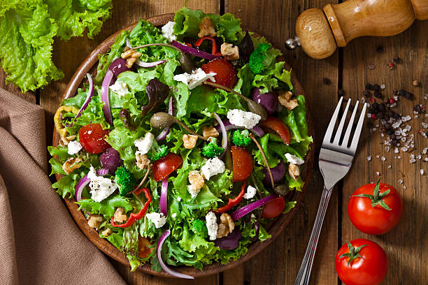 Spring salad shot from above on rustic wood table Top view of a colorful spring salad on rustic wood table. The ingredients included in the salad are lettuce, radicchio, spanish onion, black olives, capers, broccoli, bell pepper, walnut and goat cheese. The salad is served on a wood plate and a fork is beside it. A pepper grinder is tilted coming from the upper-right side of the frame and a textile napkin is at the lower-left of the frame.  DSRL studio photo taken with Canon EOS 5D Mk II and Canon EF 70-200mm f/2.8L IS II USM Telephoto Zoom Lens cheese goat cheese gourmet food stock pictures, royalty-free photos & images