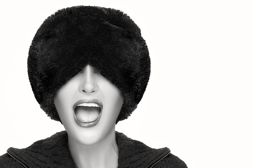 Winter beauty fashion. Close up monochrome portrait of pretty young woman with open mouth facial expression, wearing trendy fur hat that covers his eyes. Gestures and grimaces. Portrait isolated with copy space for text.