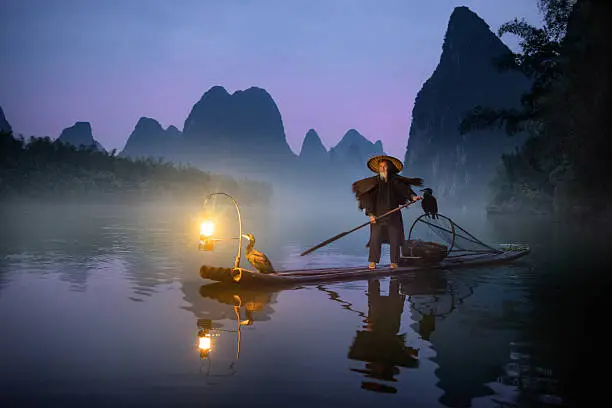 River Lee Cormorant Fisherman on bamboo raft with two cormorant birds and illuminated petrol lantern. Shot at dawn, mountains in the background.