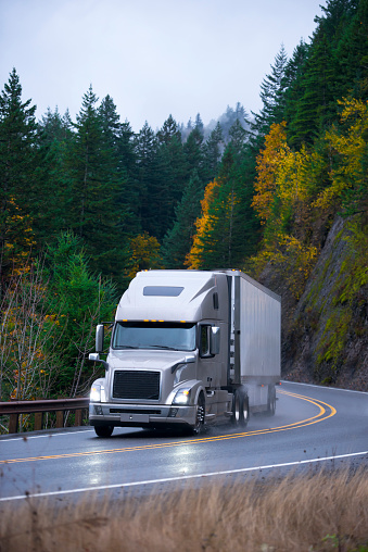 Modern professional long haul heavy semi truck with lights on and trailer for long distance transporting at the turn of the winding scenic highway with autumn trees on the rocks in the rain.