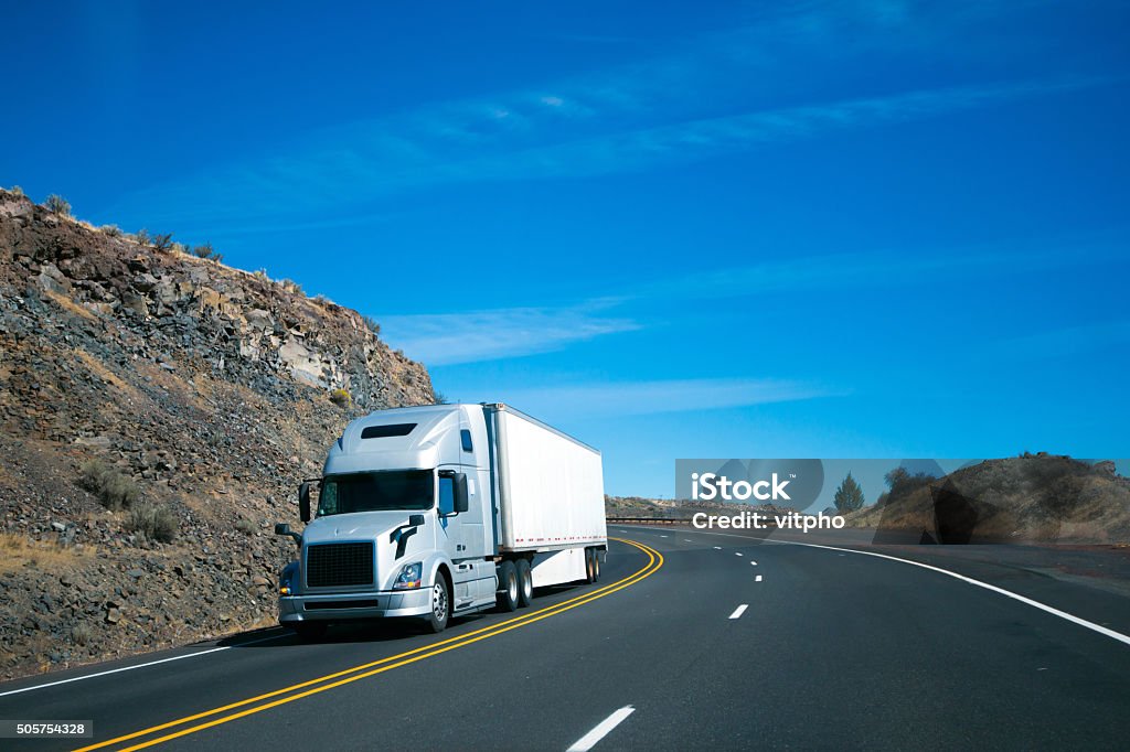 Modern semi truck and trailer on turning rocky windy road Modern long haul commercial cargo pro power silver big rig semi truck with trailer and aerodynamic to save fuel at the turn wide multilines highway among the rocky hills flanking the road. Hill Stock Photo