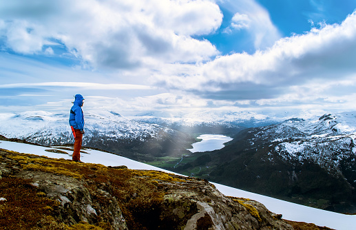 Tourist stands on a hilltop and look at the lake in the valley.  Blue sky with clouds. Snowy mountains. Sunlight breaks through the clouds. Norway.