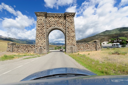 Looking down over hood of automobile while driving towards the northern arched gate at Yellowstone National Park with mountains and clouds in background.