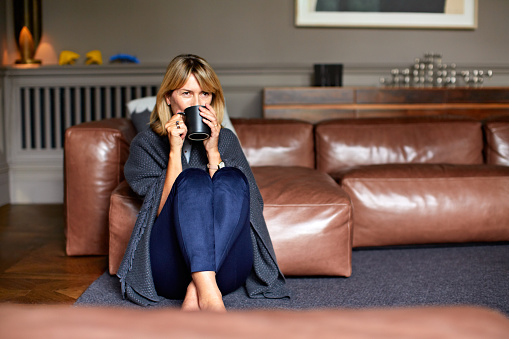 Shot of a mature woman sitting on her living room floor  drinking tea while wrapped in a blanket