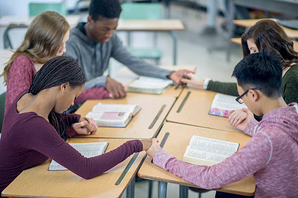 Praying Together in a Bible Study A multi-ethnic group of high school age students are sitting together at their desks and are holding hands while praying together. teenagers only teenager multi ethnic group student stock pictures, royalty-free photos & images